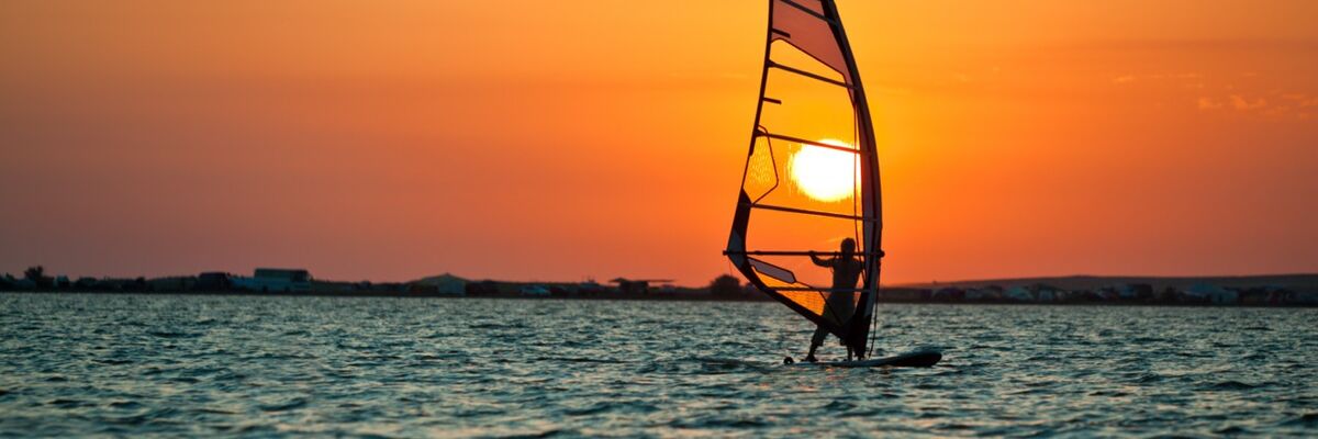 Seelandschaft mit ruhiger Wasseroberfl&auml;che, einem Windsurfer und einem goldenen Sonnenuntergang am Himmel an einem klaren Sommertag.