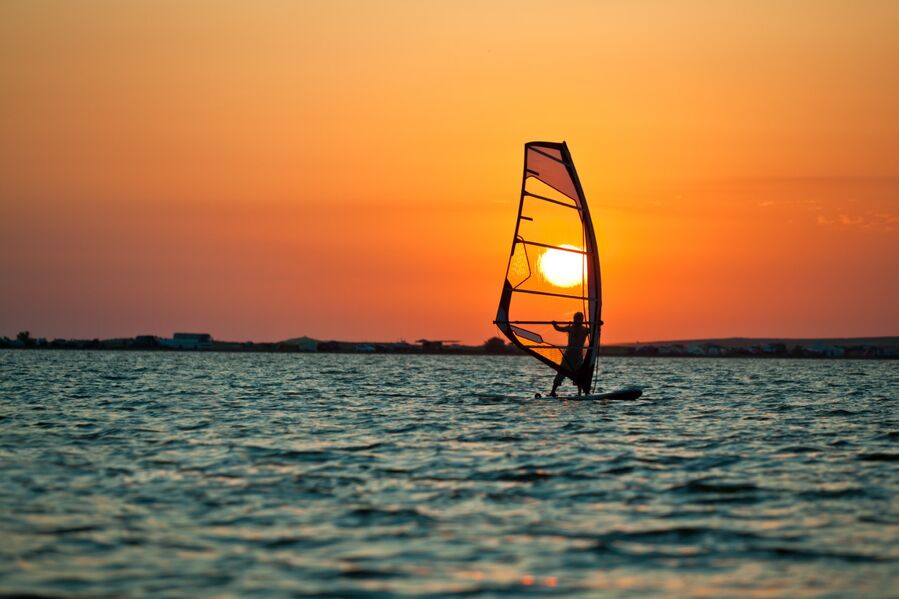 Seelandschaft mit ruhiger Wasseroberfl&auml;che, einem Windsurfer und einem goldenen Sonnenuntergang am Himmel an einem klaren Sommertag.